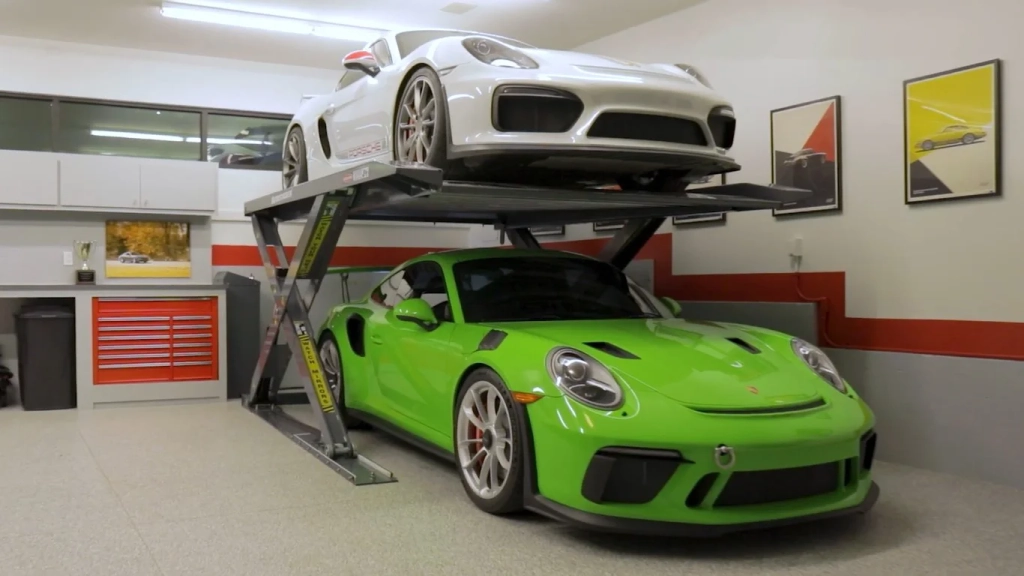 A modern home garage featuring two Porsche cars, one green and one white, parked on a hydraulic lift with the green one underneath and the white one raised above. The garage has a clean, polished floor with red and grey cabinetry, automotive-themed wall decor, and a trophy on display.
