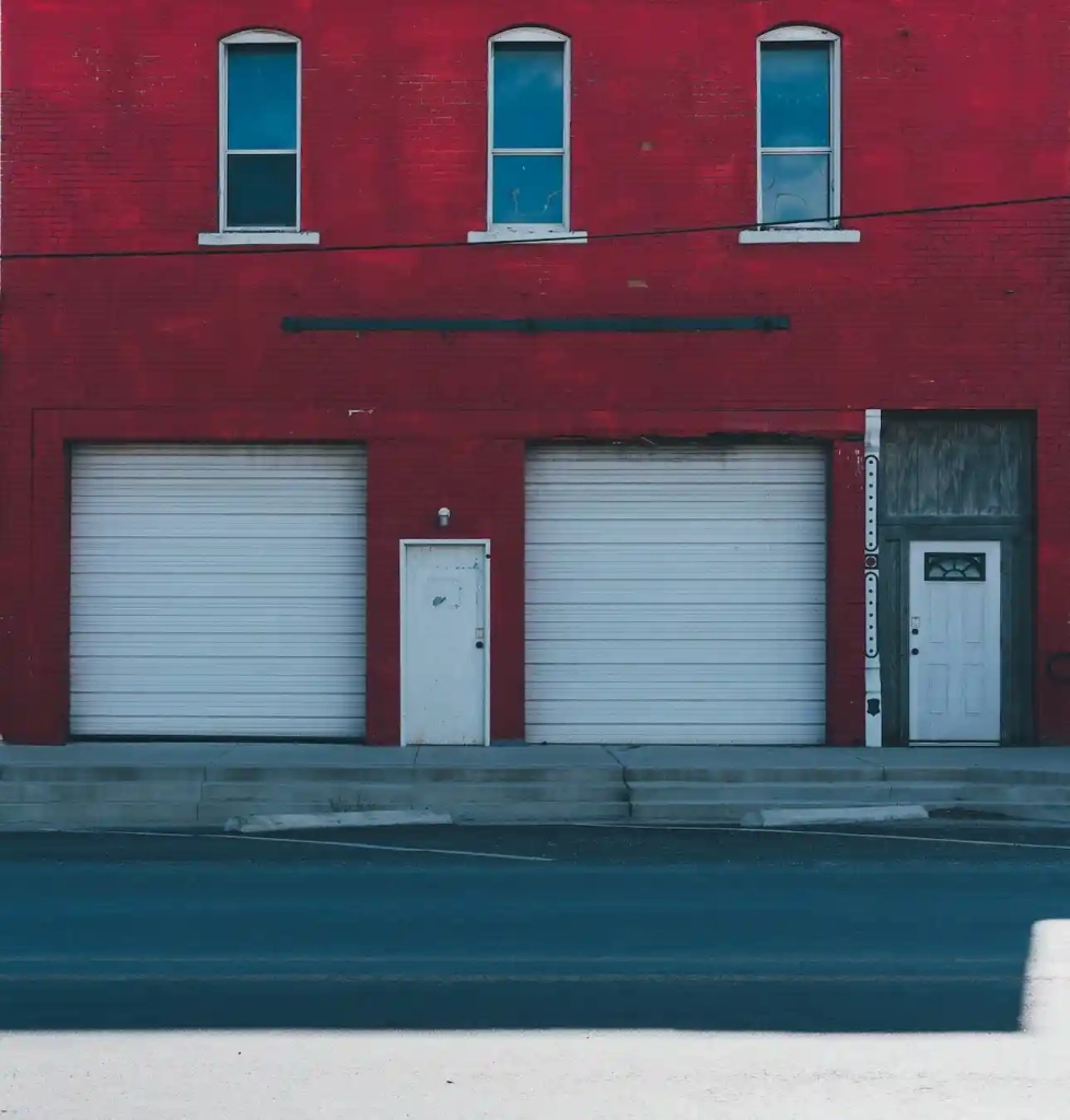 This photo depicts the front of a red brick building with two white roll-up garage doors side by side on the ground floor. There is a single white pedestrian door to the left of the garage doors and another white door to the right with a glass panel and a dark handle. Above the garage doors, there are two rectangular windows with white frames on the second story. The strong contrast between the red bricks and the white doors and windows creates a striking visual. The road in front of the building is paved and painted with a blue hue, adding to the photo's colorful aesthetic.