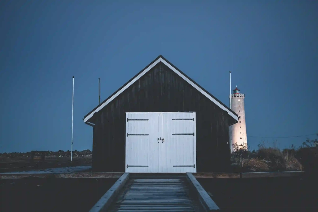 This photo captures the serene twilight scene of a dark wooden building with side-hinged double doors, painted white, in the foreground. The structure stands against a deepening blue sky. A wooden boardwalk leads up to the doors, and a flagpole stands to the left. In the background, slightly to the right, a lighthouse can be seen illuminated, providing a beacon in the dusk. The lighthouse's presence adds a maritime feel to the scene, suggesting the building may be located near the coast.