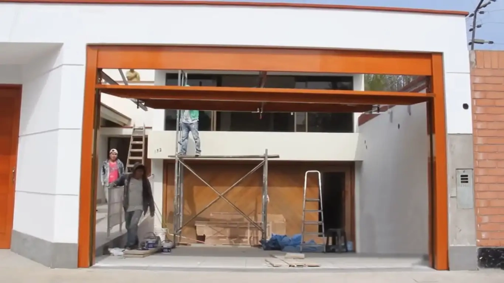 This photo shows a construction scene where workers are installing a large tilt-up overhead retractable garage door on a modern building. The door frame is a striking orange that matches the trim of the building, and the door itself is halfway open. There's a man on a scaffold inside the garage space working on the upper part of the door, while another person is walking out beneath it. The building exterior is a combination of white and orange, and there's a pedestrian door to the left of the garage door. The photo captures a moment of active construction with workers and materials present.