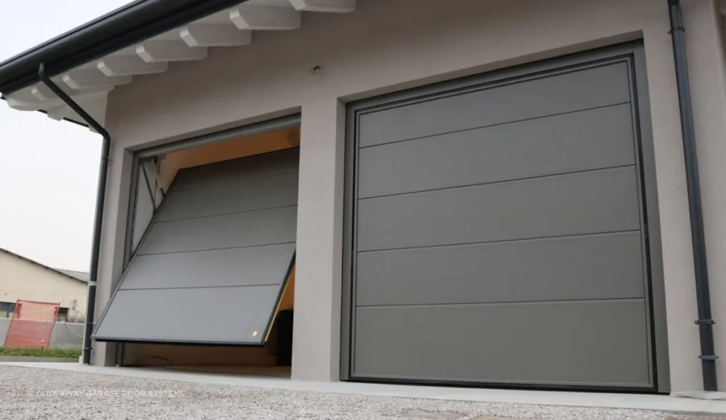 This photo depicts a modern home's garage with a tilt-up garage door that is partially open, revealing a sliver of the interior which is brightly lit. The door itself is made up of horizontal panels, painted in a muted gray that matches the home's exterior palette. Above the garage door, the eaves of the house overhang, sheltering the garage entrance. To the right of the door is a downspout attached to the exterior wall, and in the background, there appears to be an outdoor area with a fence visible. The overcast sky suggests a subdued, calm day.