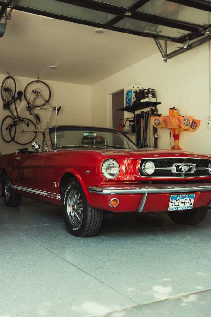 A classic red Ford Mustang convertible parked in a garage where the dust has been removed. The garage is organized with bikes mounted on the wall and shelves filled with items and sports memorabilia. The concrete floor is clean and natural light streams in through the open garage door.