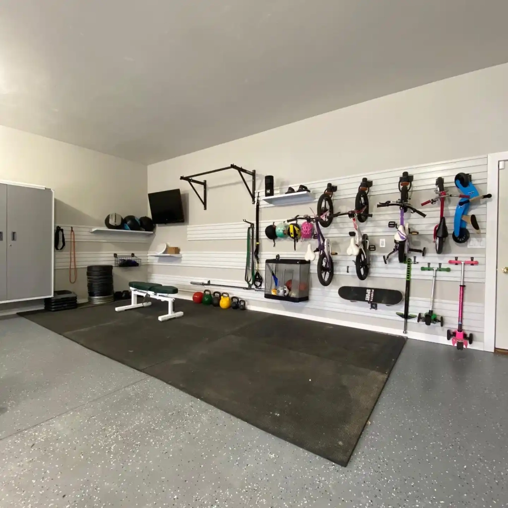 Home garage converted into a workout space with a weight bench, various exercise equipment, and bikes hung on the wall, with a large storage cabinet and a TV mounted above.